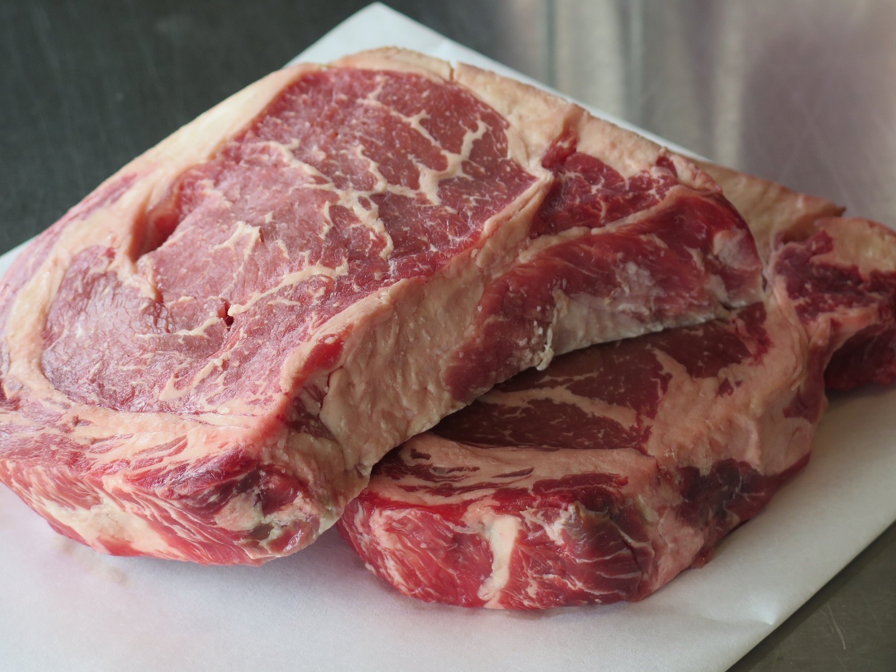 37379204_1791282624296677_4357441765733564416_n Fresh marbled ribeye steak displayed on white butcher paper, showcasing rich red meat and creamy fat content, ideal for grilling or roasting.