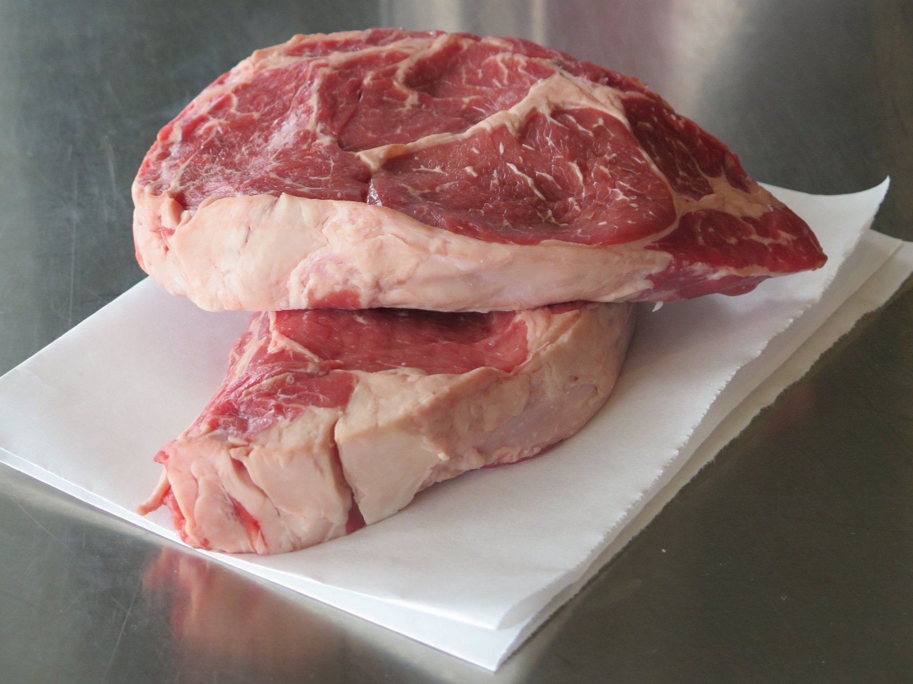 Two raw ribeye steaks placed on a white paper sheet, showcasing marbling and rich red color, ideal for grilling or pan-searing.