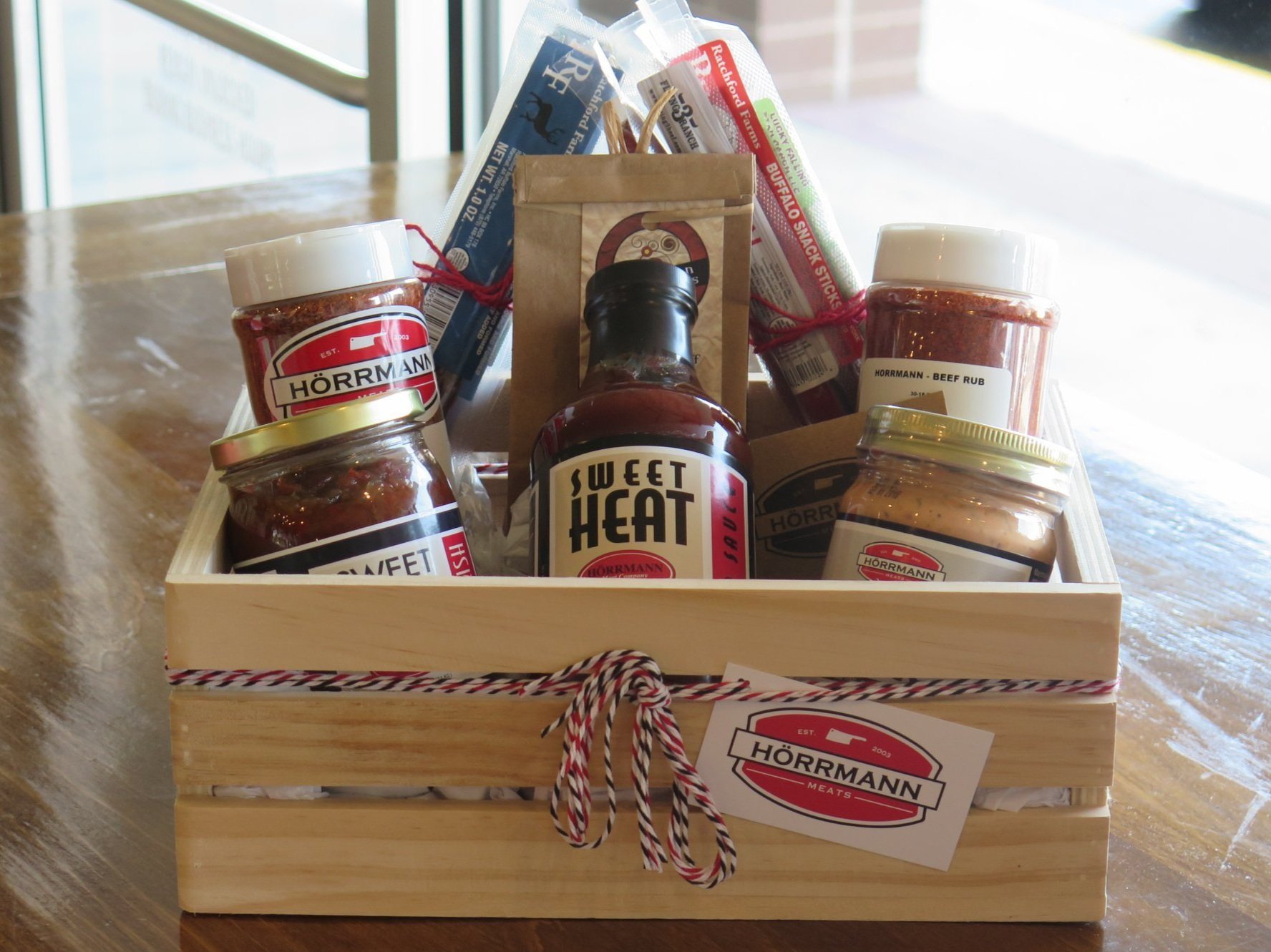 38827719_1826695174088755_3440378690834268160_n Gift basket featuring a variety of gourmet sauces and seasonings from Hörmann Meats, including Sweet Heat sauce, beef rub, and snack sticks, presented in a wooden crate with a decorative ribbon. Perfect for food lovers and grilling enthusiasts.