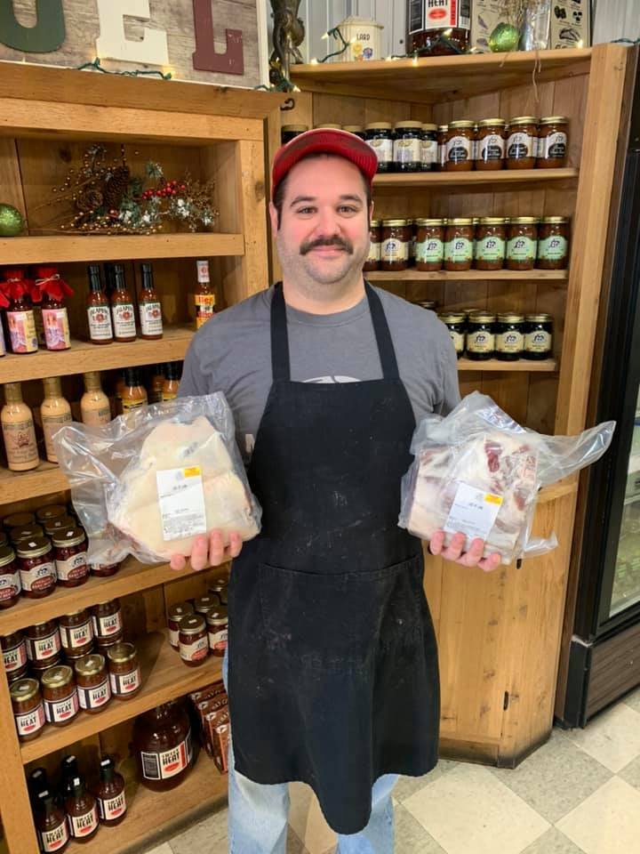 Smiling butcher wearing a black apron and red cap, holding packaged meat in each hand, standing in front of a wooden display filled with various jars and sauces.