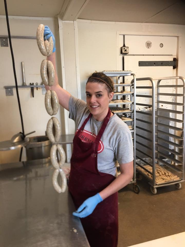 Young woman in a gray shirt and red apron holding a string of freshly made bagels in a bakery, with baking racks and kitchen equipment in the background.