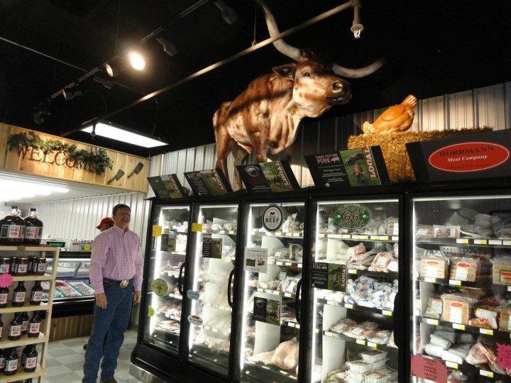 Interior of a meat market featuring a man in a pink shirt standing near refrigerated displays filled with various meat products. A decorative cow statue is mounted on the wall above the displays, with a chicken figure nearby, creating a rustic atmosphere. The store emphasizes quality meat offerings and a welcoming environment.