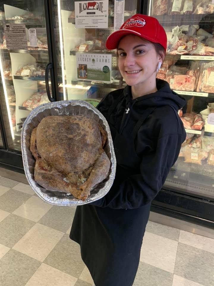 Young woman wearing a red cap and black apron smiles while holding a large, roasted turkey in a foil tray, with a meat display in the background. Ideal for promoting holiday meals and catering services.