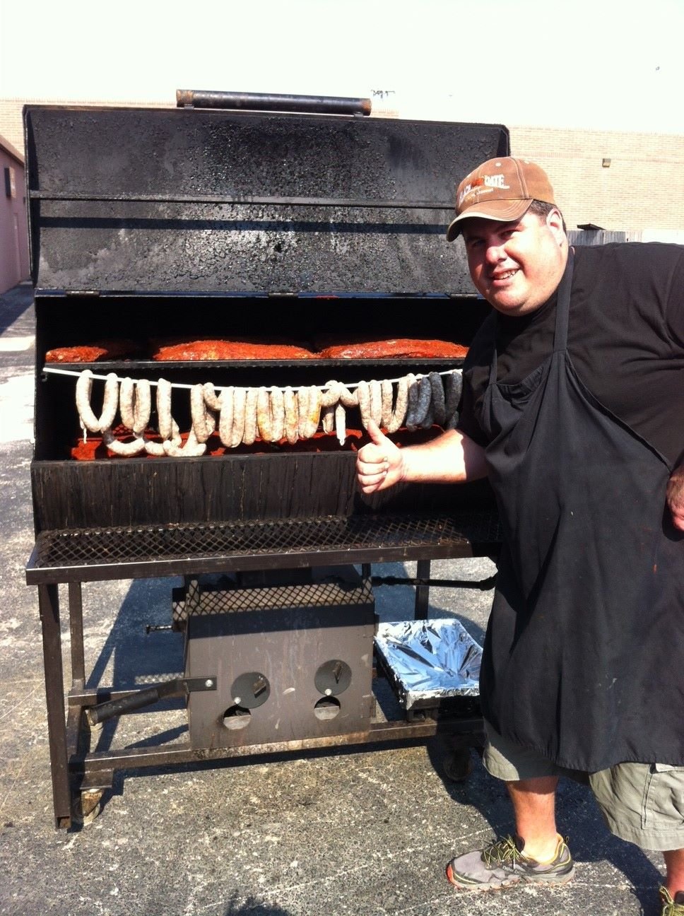 Man grilling sausages and ribs on a large outdoor barbecue smoker, giving a thumbs up in approval.